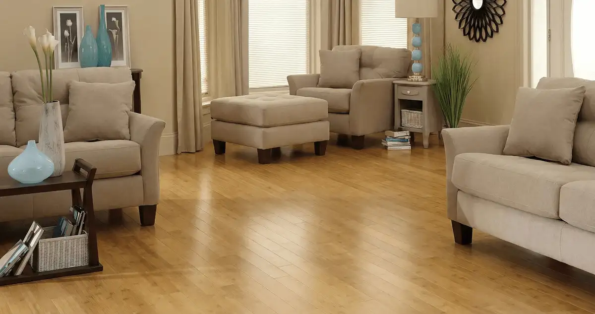 A living room with professionally installed bamboo flooring, beige sofas, a matching ottoman, a side table with a blue lamp, vases, framed pictures, and large windows with blinds and curtains in Phoenix, AZ